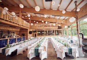 Ein geräumiger Bankettsaal mit Holzbalken, perfekt für die Hochzeitsplanung. Lange Tische sind mit weißen Tischdecken, blaugrünen Läufern und Flaschen gedeckt. Von der Decke hängen weiße Papierlaternen und Girlanden. Große Fenster und ein kleiner Balkon werten diesen wunderschönen Ort für Hochzeiten auf.