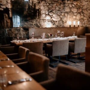 Ein gemütliches, rustikales Restaurant-Interieur mit Steinmauer und sanfter Beleuchtung bildet die perfekte Bühne für Ihre Hochzeitsfeier. Der elegant arrangierte Esstisch, komplett mit Gläsern, Tellern und einem kleinen Tafelaufsatz, ist von modernen Polsterstühlen umgeben – ein Traum für jede Hochzeitsplanung.