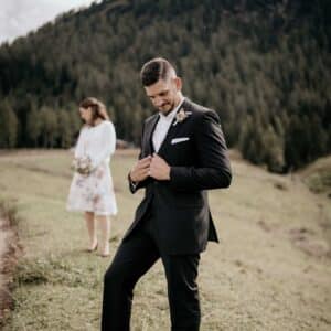 Im Vordergrund steht ein Mann im schwarzen Anzug, der im Rahmen der Hochzeitsvorbereitungen seine Jacke zurechtrückt, während hinter ihm eine Frau in einem weißen Blumenkleid mit einem Blumenstrauß steht. Sie befinden sich im Freien in einer malerischen, grasbewachsenen Gegend mit bewaldeten Hügeln.
