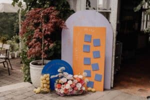 Ein Hochzeits-Sitzplan mit blauen Karten auf orangefarbenen und blauen Tafeln, mit deutschen Wörtern, umgeben von Zitronen und bunten Blumen. Eine Topfpflanze steht in der Nähe, mit einer grünen Außenansicht im Hintergrund, perfekt für diejenigen, die schon weit in ihrer Hochzeitsplanung stecken.