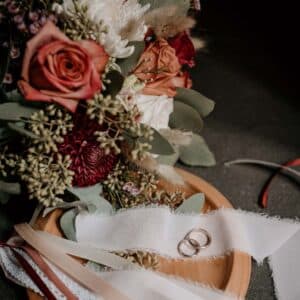 Auf einem Holztablett liegen zwei goldene Eheringe auf einem weißen Stoffstreifen und fangen damit die Essenz einer Hochzeit ein. Daneben sorgt ein Strauß Rosen und gemischter Blumen mit Grünpflanzen für rustikalen Charme. Die Szene ist vor einem dunklen Hintergrund angesiedelt, perfekt für alle, die gerade mit der Hochzeitsplanung beschäftigt sind.