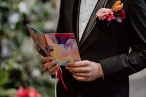 In einem frischen schwarzen Anzug und einem weißen Hemd hält die Person eine offene Karte mit farbenfrohen abstrakten Mustern in der Hand, perfekt für jede Hochzeit. Lebendige Blumen zieren ihr Revers, während der verschwommene Hintergrund auf üppiges Grün und rote Blüten hindeutet und einen eleganten Ton für diesen besonderen Anlass angibt.