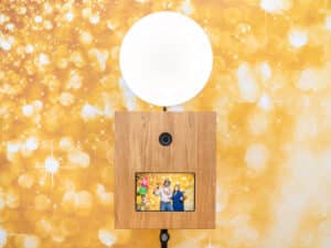 Eine Fotokabine mit Holzfassade und rundem Licht oben steht vor einem gelben, Bokeh-artigen Hintergrund mit funkelnden Lichtern, perfekt für jede Hochzeit. Der Bildschirm zeigt ein farbenfrohes Bild von drei posierenden Personen und sorgt so für Spaß bei Ihrer Hochzeitsplanung.