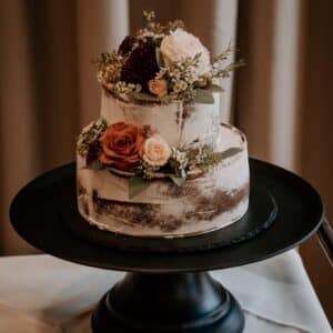 Eine zweistöckige rustikale Torte mit halbnacktem Zuckerguss-Design, perfekt für Ihre Hochzeitsplanung. Geschmückt mit Rosen und Blumen in Weiß-, Rot- und Burgundertönen wird sie auf einem schwarzen Tortenständer vor einem neutralen Hintergrund präsentiert.