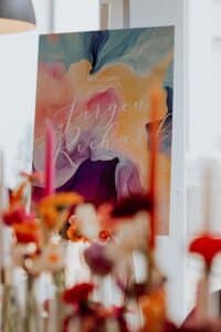 Ein farbenfrohes Willkommensschild mit abstrakten Wirbeln in verschiedenen Farbtönen wie Rosa, Blau und Gelb feiert den besonderen Anlass der Hochzeit. Der Name „Jürgen Richard“ erscheint in eleganter Handschrift, wobei der Vordergrund unscharf ist und leuchtende Blumen und Kerzen zum Vorschein bringt.