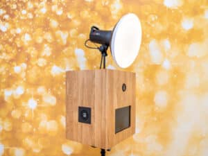 Eine moderne Fotokabine mit Holzfassade und montierter runder Studioleuchte vor einem Hintergrund mit Gold- und Bokeh-Muster, perfekt zum Festhalten wertvoller Erinnerungen an Ihre Hochzeit.