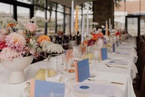 Ein langer, eleganter Esstisch ist für eine Hochzeit gedeckt. Der Tisch ist mit farbenfrohen Blumendekorationen, Kerzen, Gläsern und blauen Menükarten geschmückt. Große Fenster im Hintergrund lassen natürliches Licht herein und schaffen eine warme und einladende Atmosphäre, die perfekt ist, um die Liebe zu feiern.