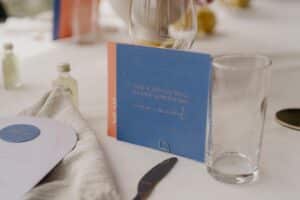 Eine Nahaufnahme eines gedeckten Tisches für eine Hochzeit zeigt eine blau-orange Karte mit deutschem Text und dem Aufdruck „anna + manuel“. Daneben liegen ein Messer, ein klares Glas, eine kleine Flasche und eine gefaltete Serviette auf der weißen Tischdecke, was die Essenz eleganter Hochzeitsplanung einfängt.