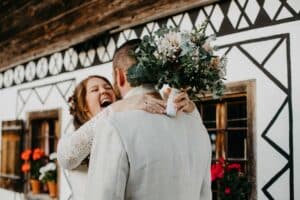 Eine Braut umarmt ihren Bräutigam freudig von hinten und hält einen Blumenstrauß in der Hand, um ihre Hochzeit zu feiern. Sie stehen vor einem rustikalen Gebäude mit weißen Wänden und geometrischen schwarzen Mustern. Leuchtend rote Blumen blühen in Blumenkästen und verleihen ihrem besonderen Tag noch mehr Charme.