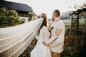 Braut und Bräutigam stehen zusammen, mit abgewandten Blicken, in einer rustikalen Umgebung im Freien. Sie trägt einen Blumenkranz und einen langen Schleier, während er einen beigen Anzug trägt. Umarmt in der Nähe eines Holzzauns mit viel Grün und einem nahe gelegenen Landhaus genießen sie die Schönheit ihres intimen Hochzeitsmoments.