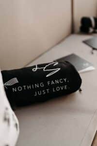Ein schwarzes Kissen mit dem weißen Text „Nothing Fancy, Just Love“ liegt auf einer hellen Oberfläche. Im Hintergrund befinden sich Notizbücher und ein verschwommenes Objekt, das Inspiration für diejenigen bietet, die tief in der Hochzeitsplanung stecken.