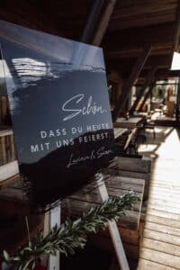 Auf einem großen Schild auf einer Staffelei steht „Schön, dass du heute mit uns feierst. Lavinia & Simon“, was dem warm beleuchteten Holzsaal Charme verleiht. Perfekt für eine Hochzeitsfeier: Eine Girlande aus Grünzeug schmückt den unteren Teil der Staffelei, während das Sonnenlicht hineinströmt und den rustikalen Innenraum erhellt.
