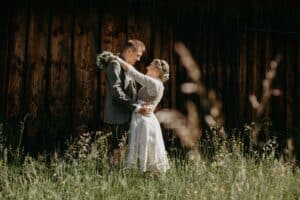 Ein Paar umarmt sich auf einer Wiese vor einem rustikalen Holzgebäude und verkörpert die Essenz einer perfekten Hochzeit. Die Frau im weißen Kleid hält einen Blumenstrauß, während der Mann einen Anzug trägt. Hohes Gras und Wildblumen umgeben sie und schaffen eine ruhige, romantische Atmosphäre, die ideal zum Heiraten ist.