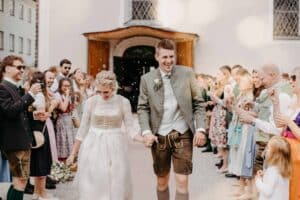 Ein Paar in traditioneller bayerischer Kleidung, die Braut im weißen Kleid und der Bräutigam in Lederhosen, lächelt, während sie Hand in Hand durch eine jubelnde Menge gehen. Gäste auf beiden Seiten klatschen und pusten Seifenblasen vor einem weißen Gebäude und feiern diese freudige Hochzeit.