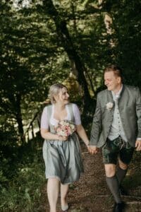 Ein Paar in traditioneller bayerischer Kleidung geht Hand in Hand einen Waldweg entlang und feiert seine Hochzeit. Die Frau trägt einen Blumenstrauß und beide lächeln, während die üppigen grünen Bäume eine ruhige Kulisse bilden.