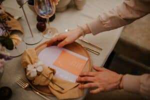 Eine Person legt ein pfirsichfarbenes Abendessenmenü auf einen Teller, der mit einer beigen Serviette und einer kleinen weißen Blume geschmückt ist. Der Tisch ist perfekt für eine Hochzeit und elegant mit Weingläsern, Besteck und sanfter Beleuchtung arrangiert, um eine einladende Atmosphäre zu schaffen.