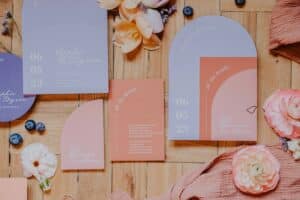 Eine Hochzeitseinladungsserie mit Karten in Pastellfarben, darunter Lavendel und Pfirsich, auf einer Holzoberfläche. Umgeben von Blumen und Blaubeeren enthält der Text Namen, ein Datum und „alle Details“ für Ihre perfekte Hochzeit.