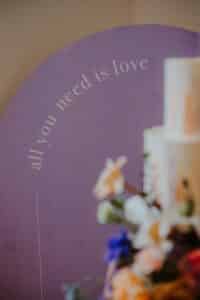 Ein verschwommenes Blumenarrangement im Vordergrund mit einem violetten Hintergrund und dem Text „All you need is love“ im Hintergrund verleiht dem Bild eine verträumte Atmosphäre, die perfekt für die Hochzeitsplanung ist.