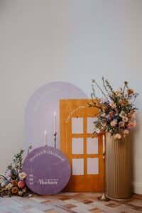 Ein Hochzeitsbegrüßungsschild besteht aus runden Schildern, auf einem steht „Herzlich Willkommen“ neben eleganten Hochzeitsdetails. Ein großes Blumenarrangement besteht aus rosa und lila Blüten, ergänzt durch drei Messingkerzenständer mit brennenden Kerzen – eine perfekte Note für Ihre Traumhochzeit.