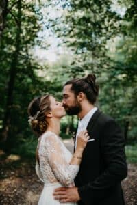 In einer ruhigen, bewaldeten Umgebung teilen Braut und Bräutigam einen intimen Moment. Sie trägt ein Spitzenkleid mit floralem Haarschmuck und er einen dunklen Anzug. Als Teil ihres wahrgewordenen Hochzeitstraums haben beide lange Haare und umarmen sich zärtlich inmitten der Bäume.