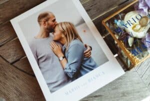 Ein Paar umarmt sich auf einem Foto, wobei der Mann die Stirn der Frau küsst. Auf dem Bild steht „Mario und Bianca“. Daneben steht eine Glasbox mit blauen Blumen und dem Namen „Mario“. Inmitten ihrer Hochzeitsplanung sind sie auf einer Holzoberfläche festgehalten und feiern die Reise der Liebe.