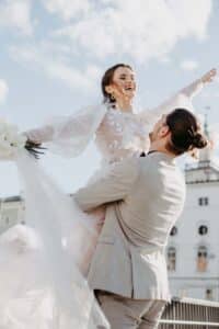 Eine lächelnde Braut in einem fließenden weißen Kleid wird freudig von einem Bräutigam in einem hellgrauen Anzug hochgehoben und fängt die Essenz ihrer Hochzeit ein. Im Freien unter einem teilweise bewölkten Himmel spiegelt die Szene perfekte Hochzeitsplanung wider, während die Braut einen Strauß weißer Blumen vor der Kulisse eines charmanten Gebäudes hält.