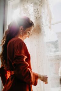 Eine Frau in einem roten Gewand steht am Fenster und lächelt sanft, während sie von ihrer bevorstehenden Hochzeit träumt. Ihr langes Haar fällt ihr über den Rücken, während das Sonnenlicht hindurchströmt und das weiße Spitzenkleid beleuchtet, das daneben hängt – und wartet auf den perfekten Moment zum Heiraten.
