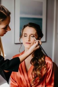 Eine Frau in einem roten Bademantel sitzt mit geschlossenen Augen da und bereitet sich auf den großen Tag vor, während eine andere Person mit einem Pinsel Make-up aufträgt. Der Hintergrund fängt die Eleganz der Hochzeitsplanung ein, wobei ein gerahmtes Kunstwerk zur Atmosphäre beiträgt.