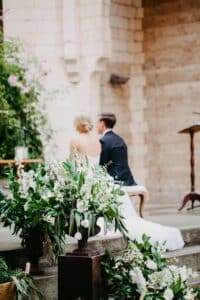 Braut und Bräutigam sitzen während ihrer Hochzeitszeremonie Seite an Seite. Sie stehen vor einer rustikalen Steinmauer, umgeben von üppigen Blumenarrangements aus weißen Blumen und Grünpflanzen. Die Szene strahlt Eleganz und Romantik aus.