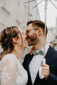 Braut und Bräutigam verbringen an ihrem Hochzeitstag einen freudigen Moment unter einem durchsichtigen Regenschirm. Die Braut trägt ein Spitzenhochzeitskleid und der Bräutigam einen dunklen Anzug mit einer grünen Fliege. Sie sind kurz davor, sich zu küssen und lächeln sich dabei an.