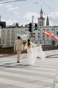 Ein Paar im Hochzeitskleid geht Hand in Hand über eine Straße, wobei die Frau ihren Schleier hält. Sie stehen vor einem historischen Gebäude mit einem Uhrenturm, daneben wehen rot-weiße Fahnen. Der Himmel ist hell und teilweise bewölkt – perfekt für diesen zauberhaften Hochzeitsmoment.