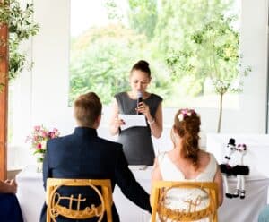 Eine Frau steht auf einem Hochzeitsempfang, hält ein Mikrofon und liest aus einer Zeitung vor. Ihr gegenüber sitzt ein Paar mit dekorierten Stühlen, auf denen „besser“ und „zusammen“ steht. Ein Blumenarrangement und ausgestopfte Pinguine verleihen dem hellen Raum Charme.