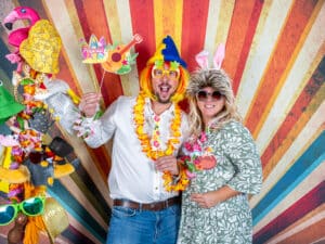 Ein Mann und eine Frau posieren verspielt vor einem bunten, gestreiften Hintergrund. Mit lustigen Accessoires wie Hüten und Sonnenbrillen verkörpern sie den hawaiianischen Geist. Der Mann hält ein „Hawaii“-Schild und verleiht ihrer Hochzeitsplanung eine tropische Note. Beide lächeln und genießen den Moment.