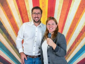Ein lächelnder Mann und eine lächelnde Frau stehen vor einem bunten, gestreiften Hintergrund, als würden sie „Ja, ich will“ sagen. Die Frau, gekleidet in einen grauen Blazer, hält ein Mikrofon, als würde sie ihre Hochzeitspläne verkünden, während der Mann ein weißes Hemd und Jeans trägt. Beide blicken glücklich in die Kamera.