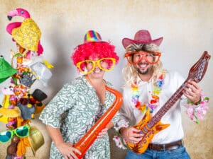 Zwei Personen in farbenfrohen Kostümen posieren mit übergroßen Sonnenbrillen und Perücken und verleihen dieser Hochzeitsfeier eine verspielte Note. Einer hält ein Spielzeugsaxophon, der andere eine Spielzeuggitarre. Daneben steht ein Ständer mit Hüten und Masken, der die festliche Stimmung einfängt und perfekt für eine innovative Hochzeitsplanung ist.