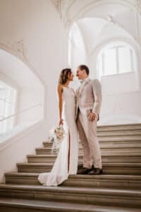 Braut und Bräutigam stehen auf einer eleganten Treppe und blicken einander an. Die Braut trägt ein weißes Kleid mit einem Blumenstrauß, während der Bräutigam einen hellen Anzug trägt. Diese atemberaubende Hochzeitskulisse zeichnet sich durch kunstvolle Bögen und große Fenster aus, perfekt für diejenigen, die ihre Hochzeitsplanung gewissenhaft durchführen.