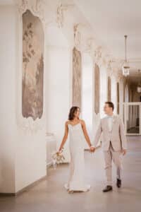 Eine Braut im weißen Kleid und ein Bräutigam im hellgrauen Anzug gehen Hand in Hand einen eleganten Flur entlang, der an ihre sorgfältige Hochzeitsplanung erinnert. Der Raum ist mit gerahmter Wandkunst und kunstvollen Zierleisten geschmückt. Sie hält einen Blumenstrauß und beide lächeln sich an.