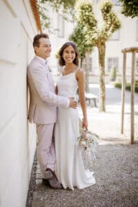 Eine Braut in einem weißen Kleid und ein Bräutigam in einem hellen Anzug stehen lächelnd und umarmend vor einer sonnenbeschienenen Wand und fangen ihre Hochzeitsfreude ein. Die Braut hält einen kleinen Blumenstrauß, im Hintergrund sind Bäume und ein Gebäude zu sehen.