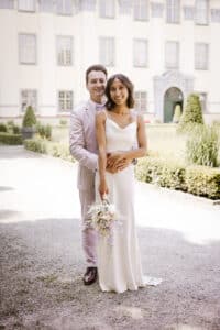 Ein lächelndes Paar steht draußen vor einem prachtvollen Gebäude. Die Frau, strahlend in einem weißen Kleid und mit einem Blumenstrauß in der Hand, sieht an ihrem Hochzeitstag aus wie das Bild der Eleganz. Der Mann, elegant in einem hellen Anzug, legt liebevoll seine Arme um sie unter dem sonnigen Himmel – ein perfekter Moment, geboren aus sorgfältiger Hochzeitsplanung.