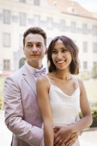 In einer sonnenbeschienenen Umgebung im Freien steht ein Paar eng beieinander im Hochzeitskleid und genießt die Freude der Hochzeit. Der Mann trägt einen hellrosa Anzug mit Fliege, während die Frau in ihrem weißen ärmellosen Kleid strahlt und seine Hand hält. Im Hintergrund ist ein Gebäude leicht verschwommen.