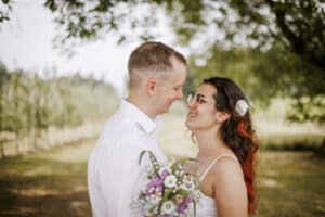 Ein Paar steht sich draußen unter einem Baum gegenüber und feiert seine Hochzeit. Die Frau, die einen Strauß Wildblumen in der Hand hält und eine Sonnenbrille trägt, lächelt den Mann im weißen Hemd herzlich an. Ihre Verbindung ist so natürlich und schön wie die Landschaft, die sie umgibt.