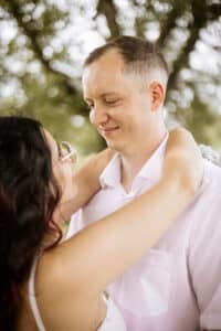 Ein Paar umarmt sich im Freien; die Frau blickt abgewandt, während der Mann sie anlächelt und einen Moment der Liebe auf ihrer Hochzeitsreise einfängt. Sie hat dunkles Haar und eine Sonnenbrille, er hat kurze Haare und trägt ein weißes Hemd. Im Hintergrund sind grüne, verschwommene Bäume zu sehen, die eine Parkkulisse suggerieren.