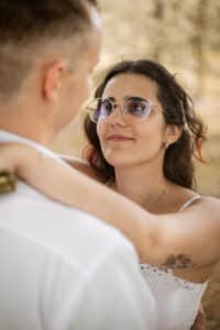 Eine Frau mit Brille und weißem Kleid, die sich möglicherweise ihre Hochzeit vorstellt, blickt zu der Person vor ihr im Freien auf. Mit den Armen, die liebevoll um ihren Hals geschlungen sind, verleiht ein sanft verschwommener Hintergrund eine Atmosphäre der Intimität und Freude.