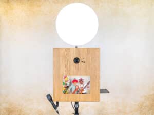 Eine charmante Vintage-Fotokabine aus Holz, perfekt für die Hochzeitsplanung, verfügt über ein rundes LED-Licht oben, ein Kameraobjektiv in der Mitte und einen kleinen Bildschirm, der ein farbenfrohes Bild anzeigt. Am Ständer ist ein Mikrofon befestigt, das vor einer strukturierten beigen Wand steht.