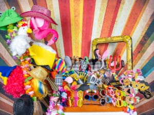 Eine lebendige Sammlung farbenfroher Kostümrequisiten vor einem bunten, strahlenden Hintergrund. Zu den Artikeln gehören Hüte, Brillen, Perücken, Masken, ein Flamingo und mehr – perfekt, um jeder Hochzeitsfotokabine während Ihrer Hochzeitsplanung Flair zu verleihen.