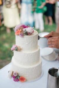 Eine mehrstöckige weiße Hochzeitstorte, geschmückt mit bunten Blumen, wird angeschnitten. Die Torte steht auf einer weißen Tischdecke und ist mit Gerbera und Rosen geschmückt. Im Hintergrund tummeln sich verschwommene Gäste auf einer Rasenfläche und tragen zur fröhlichen Atmosphäre dieser unvergesslichen Hochzeitsfeier bei.