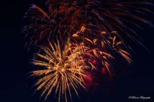 Ein farbenprächtiges Feuerwerk erhellt den Nachthimmel. Goldene Funken schießen in dichten Büscheln nach außen, einige davon mit violetten Schattierungen, und spiegeln die Freude einer Hochzeitsfeier wider. Die farbenfrohen Explosionen vor dem dunklen Hintergrund spiegeln die Aufregung und Pracht der Hochzeitsfeierlichkeiten wider.