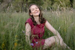 Eine Frau in einem rosa Kleid sitzt lächelnd auf einem Feld mit hohem Gras und Wildblumen, umgeben von Grün – eine perfekte Kulisse, die Natur mit der Freude am Heiraten verbindet. Im Hintergrund sind verschwommene Bäume zu sehen, die diese friedliche und romantische Szene noch verstärken.