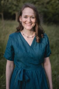 Eine Person mit langen braunen Haaren steht lächelnd im Freien und trägt ein blaues Wickelkleid – perfekt für einen Tag der Hochzeitsplanung. Im Hintergrund sind Grünflächen und verschwommene Bäume zu sehen, die eine bezaubernde Atmosphäre schaffen, die zu jeder Hochzeit passt.