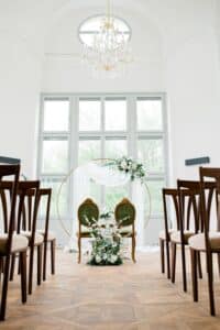 Eine helle, elegante Hochzeitszeremonie besteht aus einem runden Blumenbogen und zwei grünen Stühlen am Altar. Reihen von Holzstühlen säumen den Gang in einem geräumigen Raum mit großen Fenstern und einem Kronleuchter. Weiße Blumen und Grünpflanzen schmücken die Szene, perfekt für diejenigen, die ihre Traumhochzeit planen.